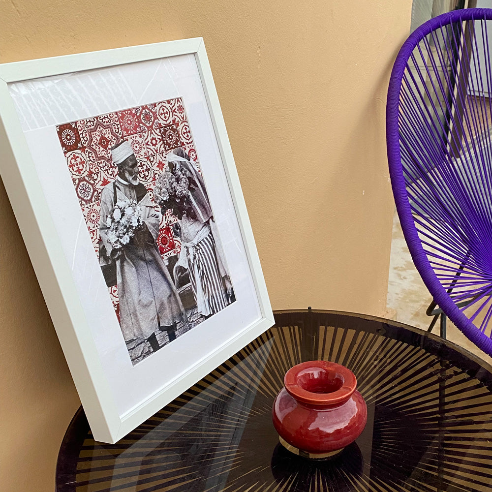 Tableau décoratif représentant un vieux couple marocain portant deux bouquets de fleurs, sur un fond de zellige marocain rouges, encadré dans un cadre blanc - Format A3.